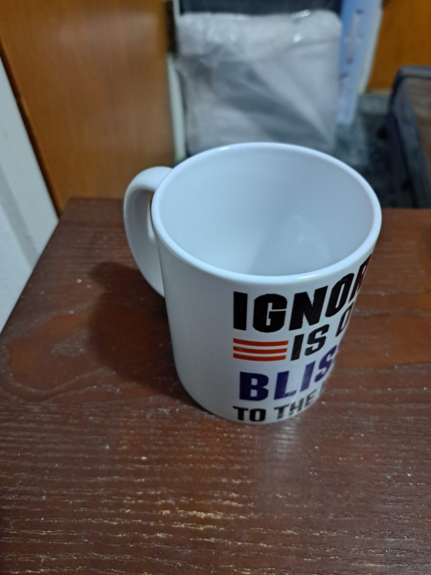 White Ignorance is Bliss Coffee Mug by Fantasy Tees Plus with bold black and blue text on wood surface